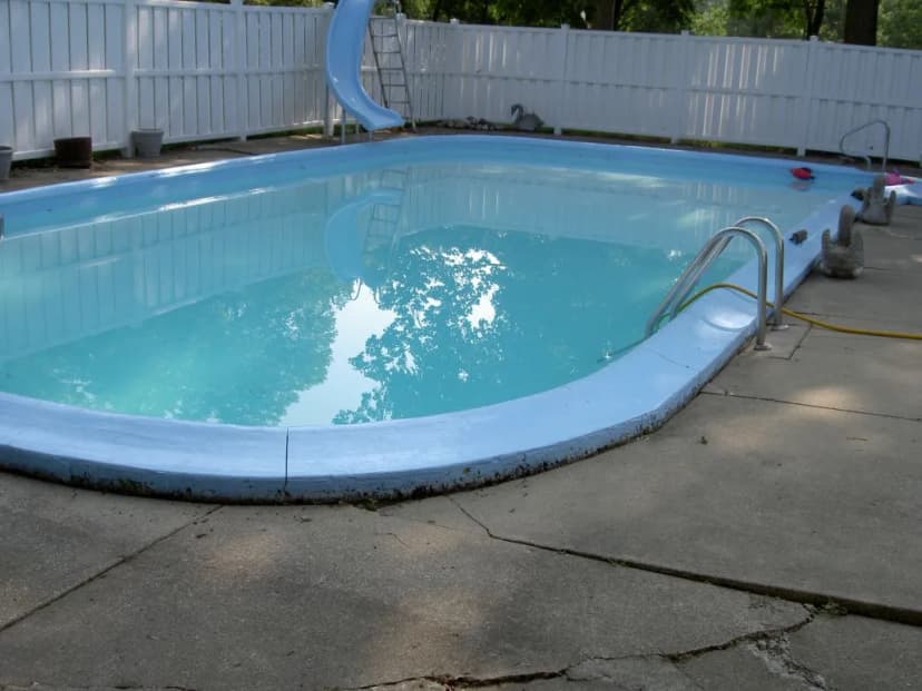 A clear blue swimming pool with a slide, surrounded by concrete and greenery.